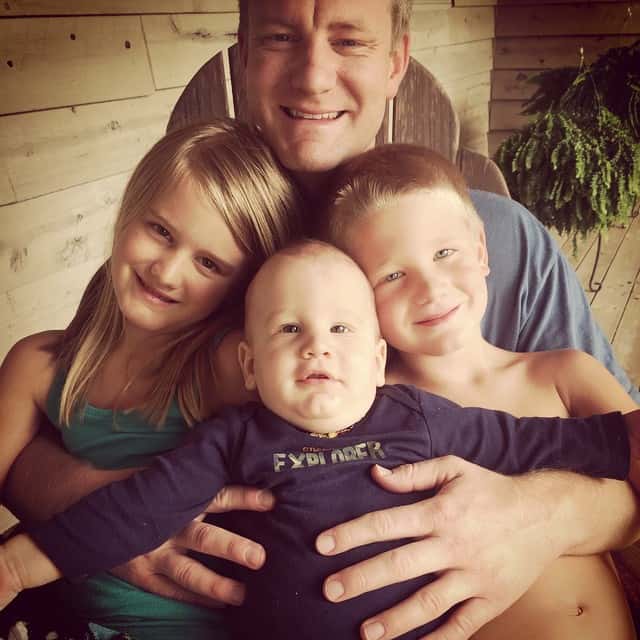 Family PIcture of a dad holding his 3 children on a front porch in a rocking chair. A green fern is behind them.