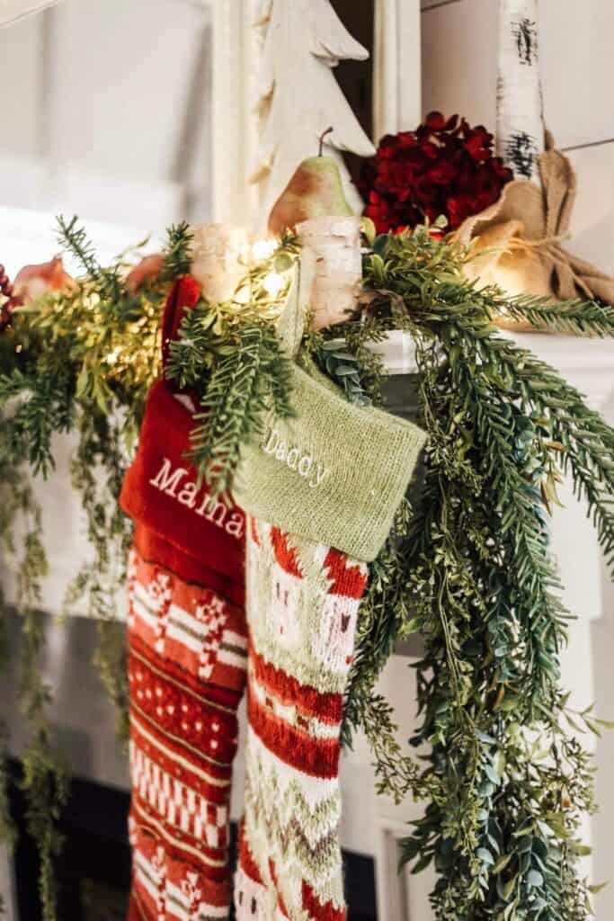 Festive holiday stockings hanging on a Christmas mantel decorated with greenery and string lights.