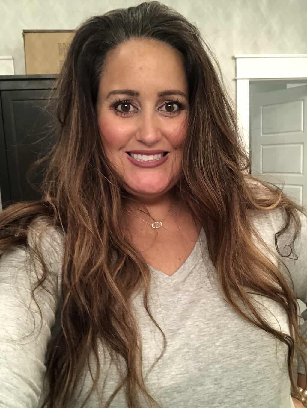 Picture of woman with long brown hair and makeup on. Her eyelashes look amazing and are curled.  She is standing up in a bathroom and has a grey shirt on and a Kendra Scott necklace.