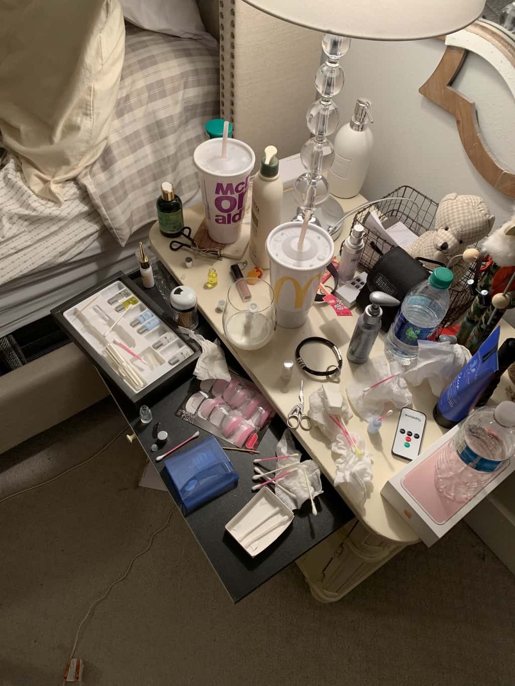 A table full of junk. Husbands side of the bed. 2 Mcdonalds cups, a wine glass with milk and a syringe, q-tips, eyelash kit, wet wipes, a bottle of water, lotion, and another bottle of lotions, and a remote and a big mess.
