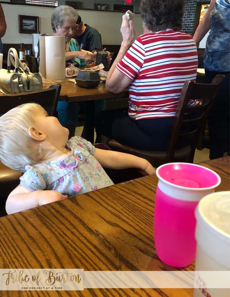 Baby in a highchair at a restaurant. Ladies at a table behind her. Pink sippy cup on table.