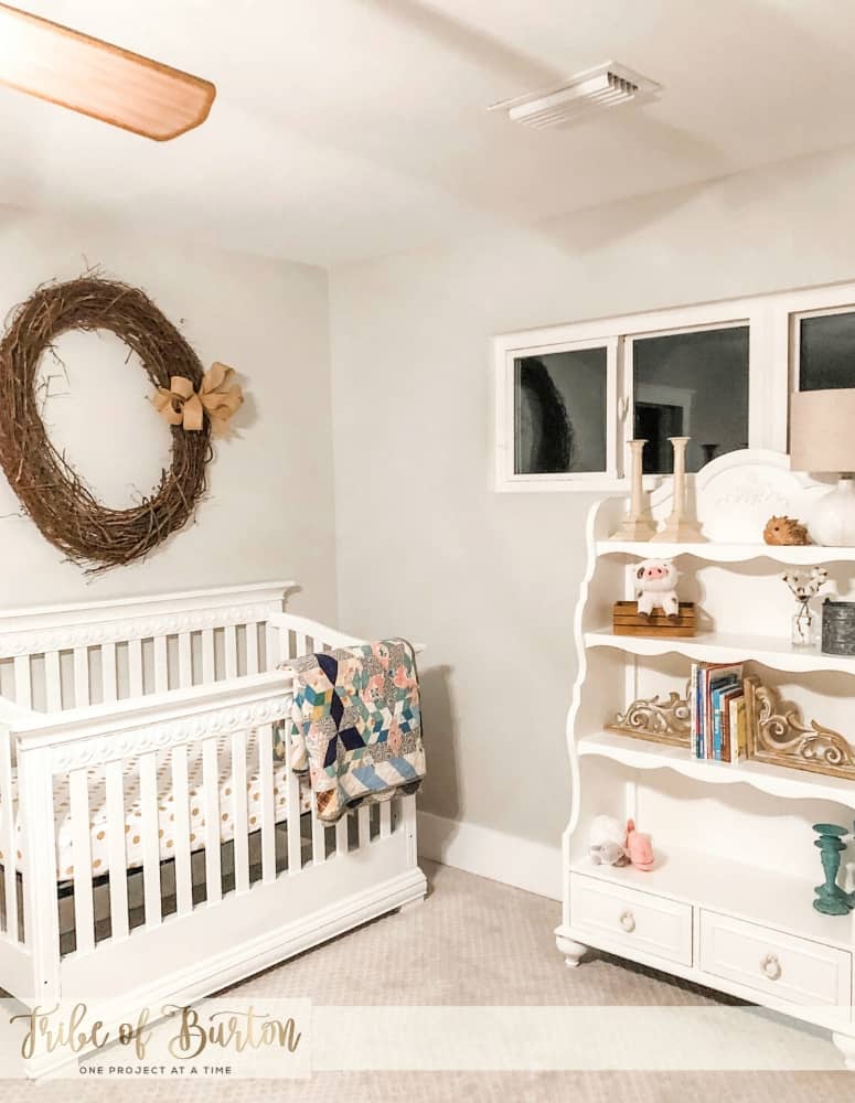 A baby crib and old quit with a wreath on the wall and a bookshelf.