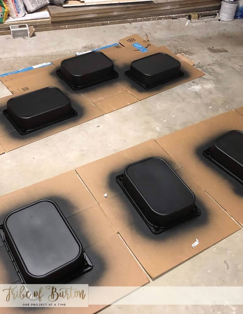 Black plastic painted buckets laying upside down on cardboard drying in a garage.