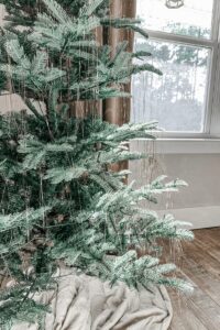 Frosted artificial Christmas tree with hanging silver ornaments and a cozy beige blanket underneath.