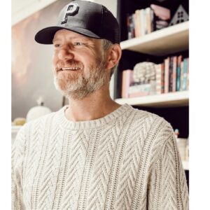 Bearded man in casual sweater and cap smiling indoors, bookshelf background, promoting community and lifestyle at Tribe of Burton.