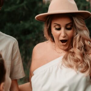 Excited woman wearing a beige hat and white top, enjoying outdoor gathering with tribe of Burton.